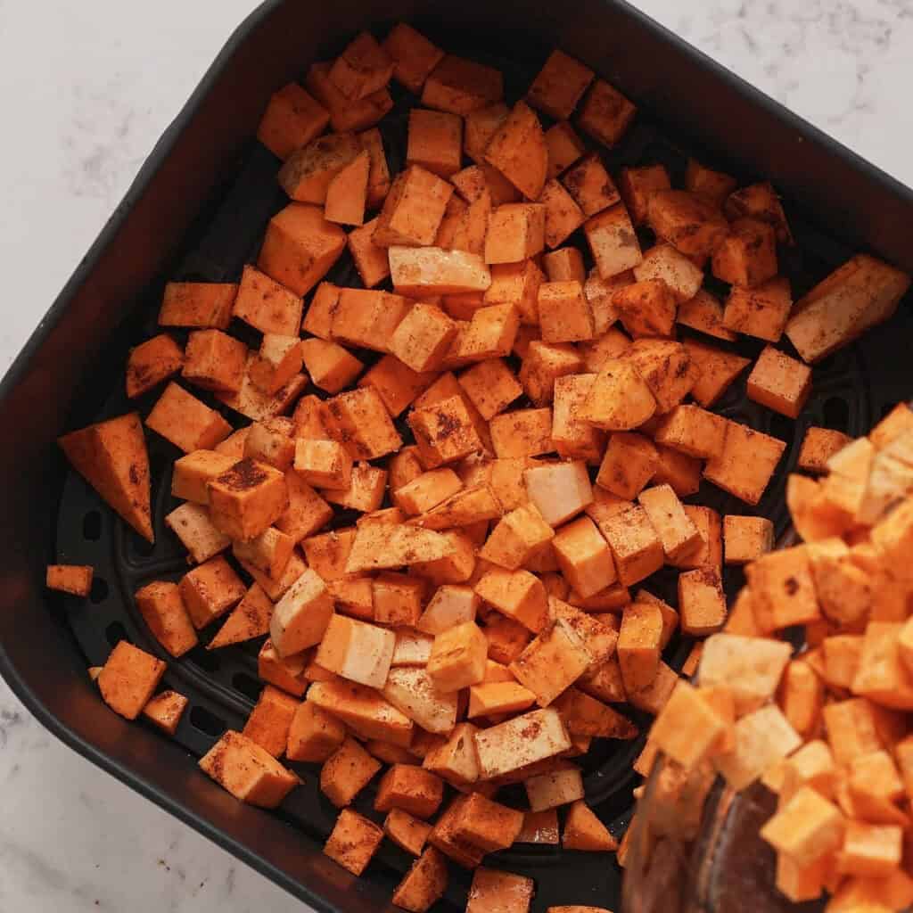 Diced sweet potatoes seasoned with spices are spread evenly in an air fryer basket, ready to be cooked. The basket sits on a marble countertop.