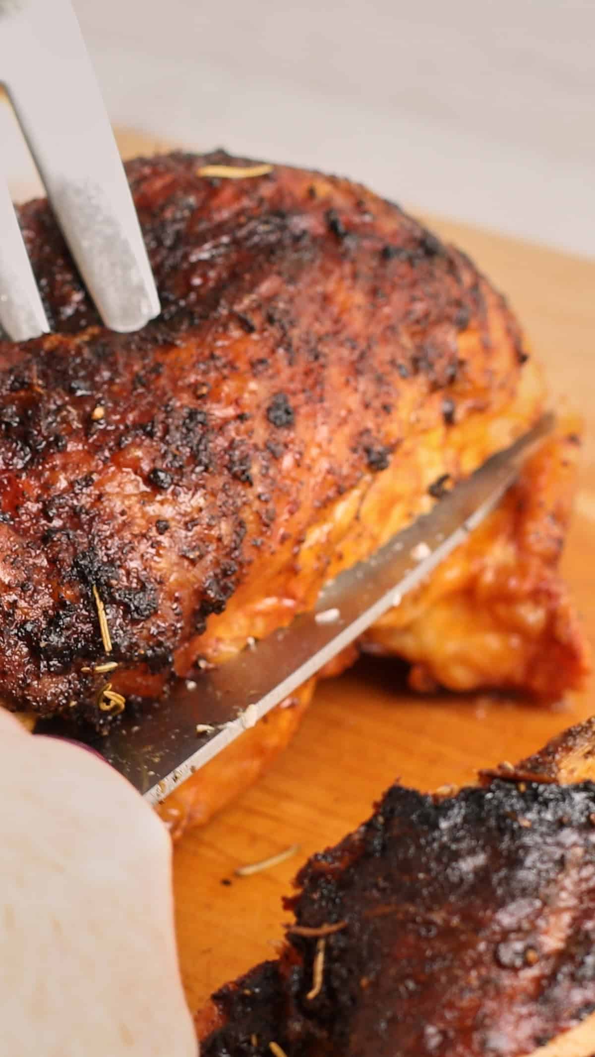 A close-up of a person slicing a juicy, seasoned air fryer turkey breast with a knife on a wooden cutting board. A fork holds the turkey breast in place as it is being cut.