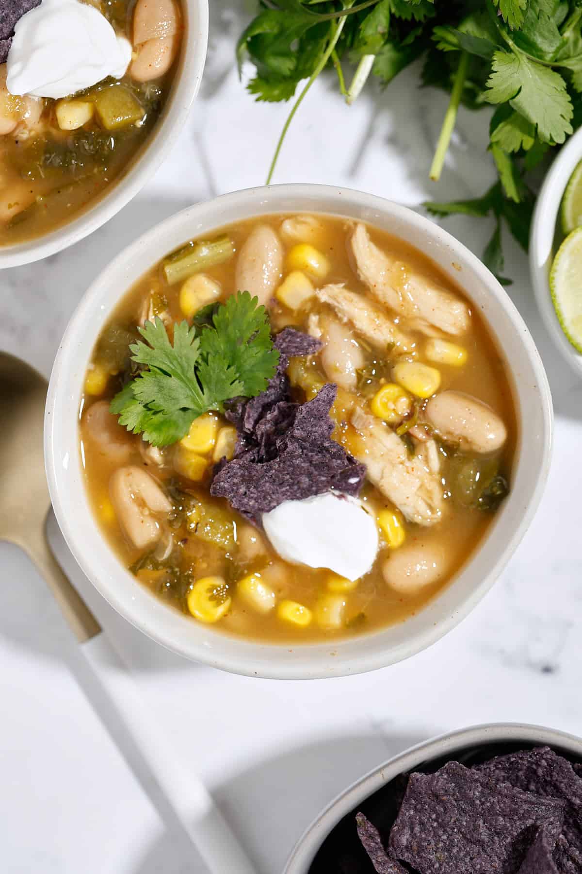 A bowl of white chicken chili with corn, white beans, shredded chicken, green chiles, topped with blue corn tortilla chips, a dollop of sour cream, and cilantro, placed on a white surface.