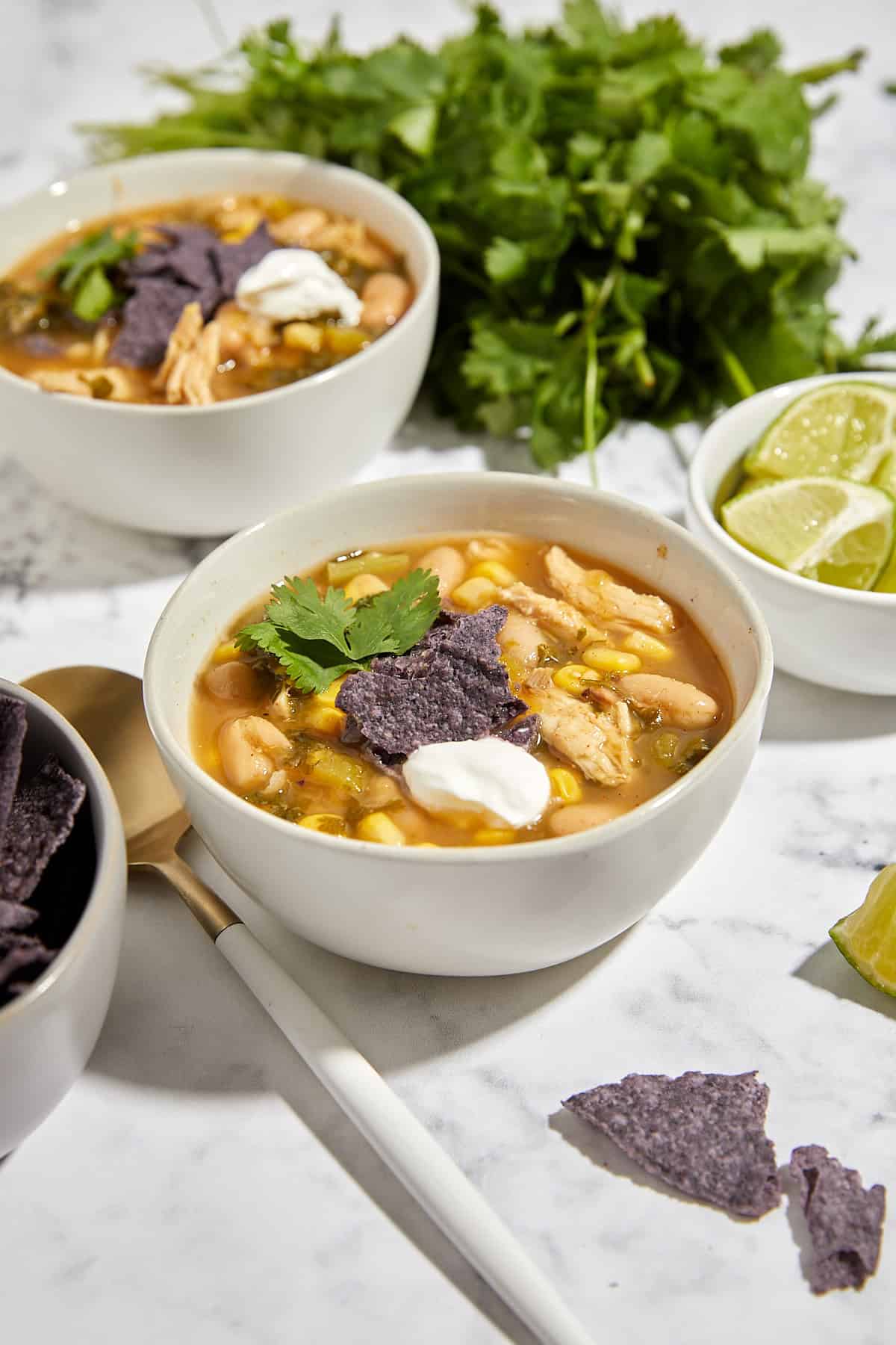 Two bowls of turkey verde tortilla soup topped with sour cream, cilantro, and blue corn tortilla chips, set on a marble surface with lime wedges and fresh cilantro in the background.
