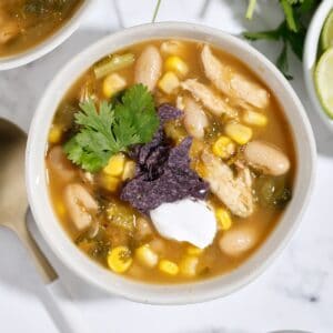 A bowl of white chicken chili topped with sour cream, blue corn tortilla chips, and fresh cilantro, featuring white beans, corn, and shredded chicken. A spoon and lime wedges are nearby on a white surface.