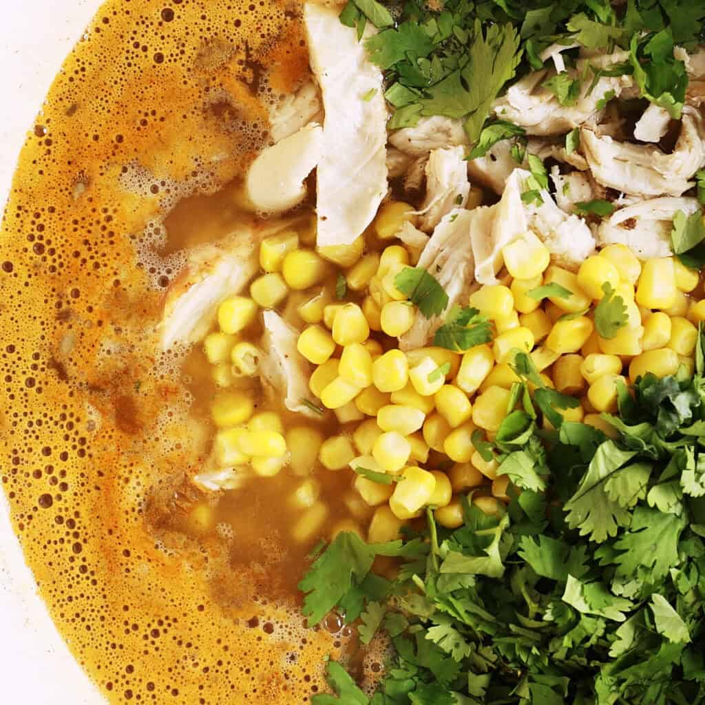 A close-up of a soup with shredded turkey, corn kernels, chopped cilantro, and a verde (green tomatillo) broth seen from above.