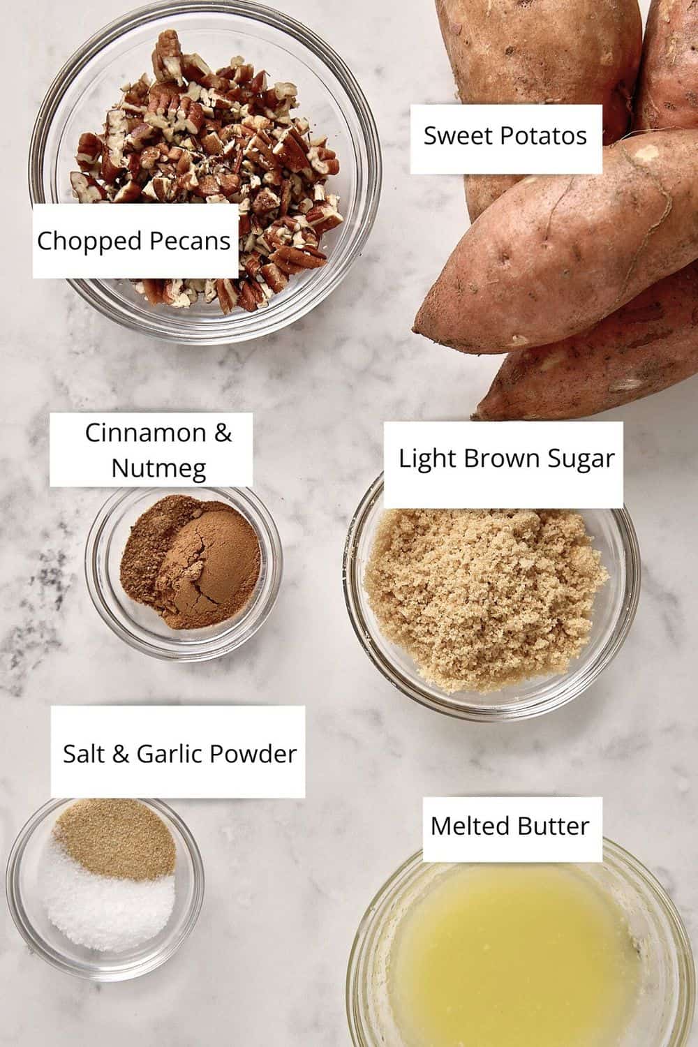 Bowls with chopped pecans, spices, light brown sugar, salt, and butter sit beside three whole sweet potatoes, perfect for making air fryer sweet potatoes cubes on a marble surface. Each ingredient is labeled.