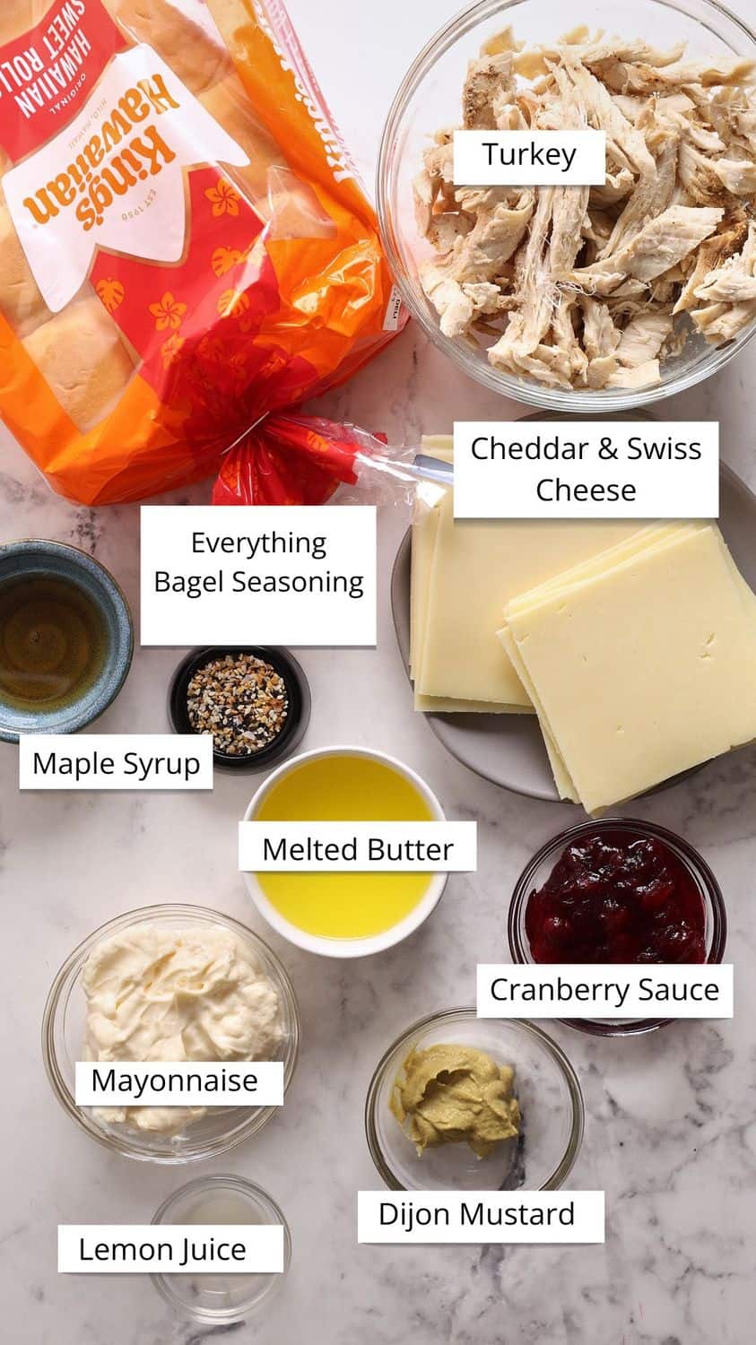 Ingredients for a turkey sandwich are arranged on a counter, including shredded turkey, cheddar and Swiss cheese, cranberry sauce, mayonnaise, Dijon mustard, lemon juice, melted butter, everything bagel seasoning, maple syrup, and buns.