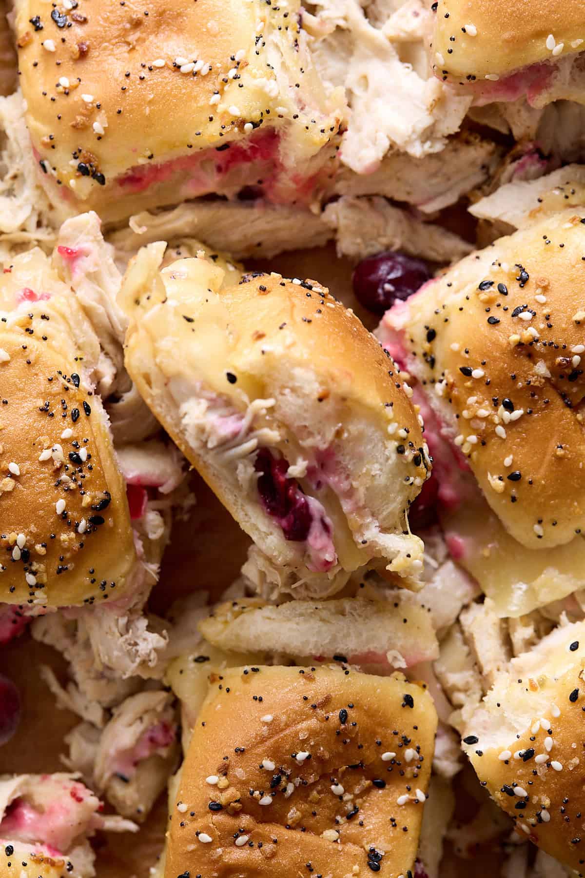 Close-up of mini sandwiches with shredded turkey, melted cheese, cranberry sauce, and poppy seed-topped buns. The filling is slightly spilling out, showing the mix of textures and colors.
