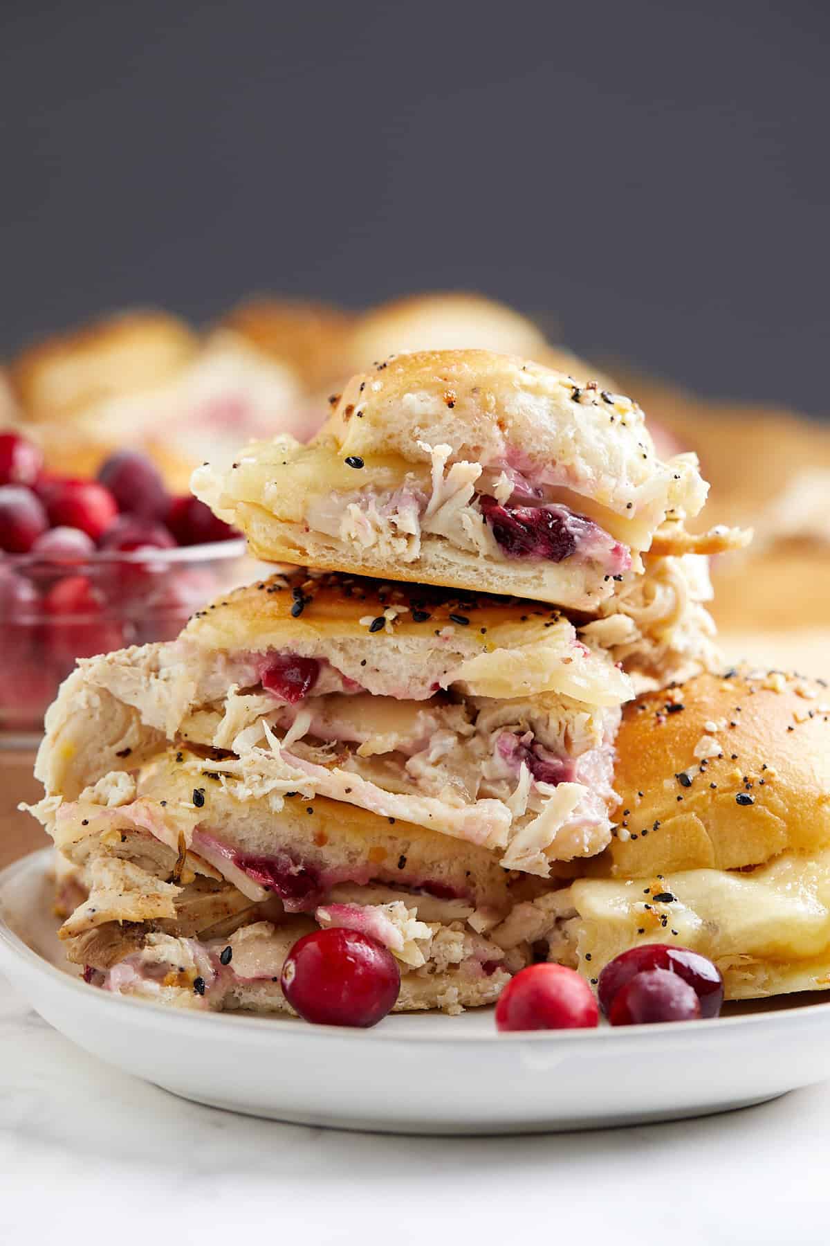 A close-up of stacked turkey sliders filled with melted cheese and cranberries on a white plate, with a few fresh cranberries scattered around. A bowl of cranberries and more sandwiches are blurred in the background.