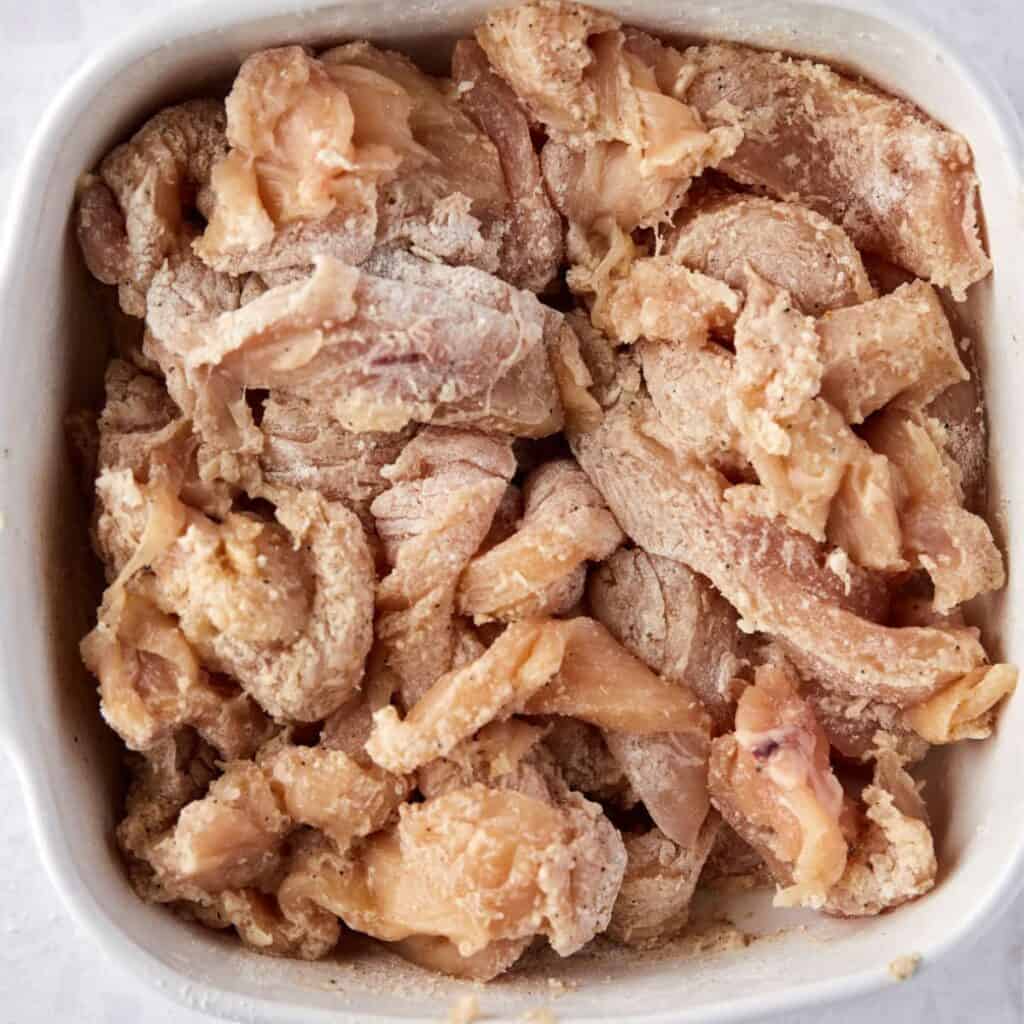 Raw, sliced chicken pieces coated in a light flour or starch mixture are piled in a white dish, ready for cooking or marinating.
