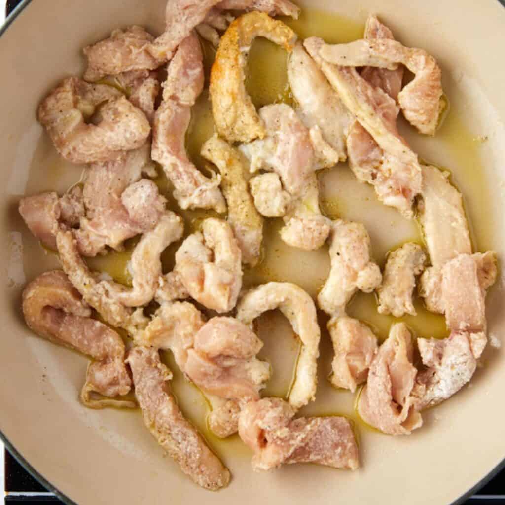 Sliced raw chicken pieces coated in seasoning are spread out in a pan with a small amount of oil, ready to be cooked.