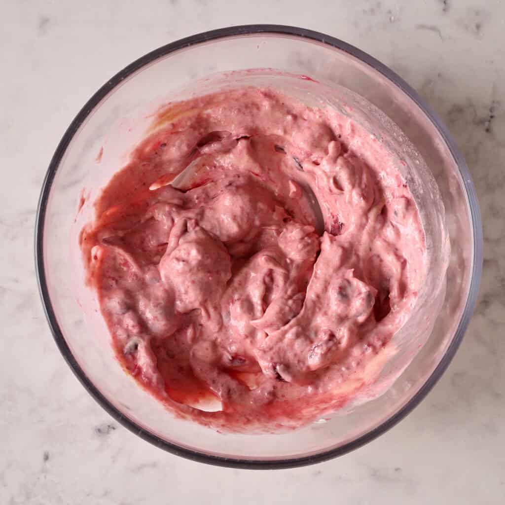 A glass bowl on a marble surface containing thick, pink batter with visible fruit chunks mixed in.