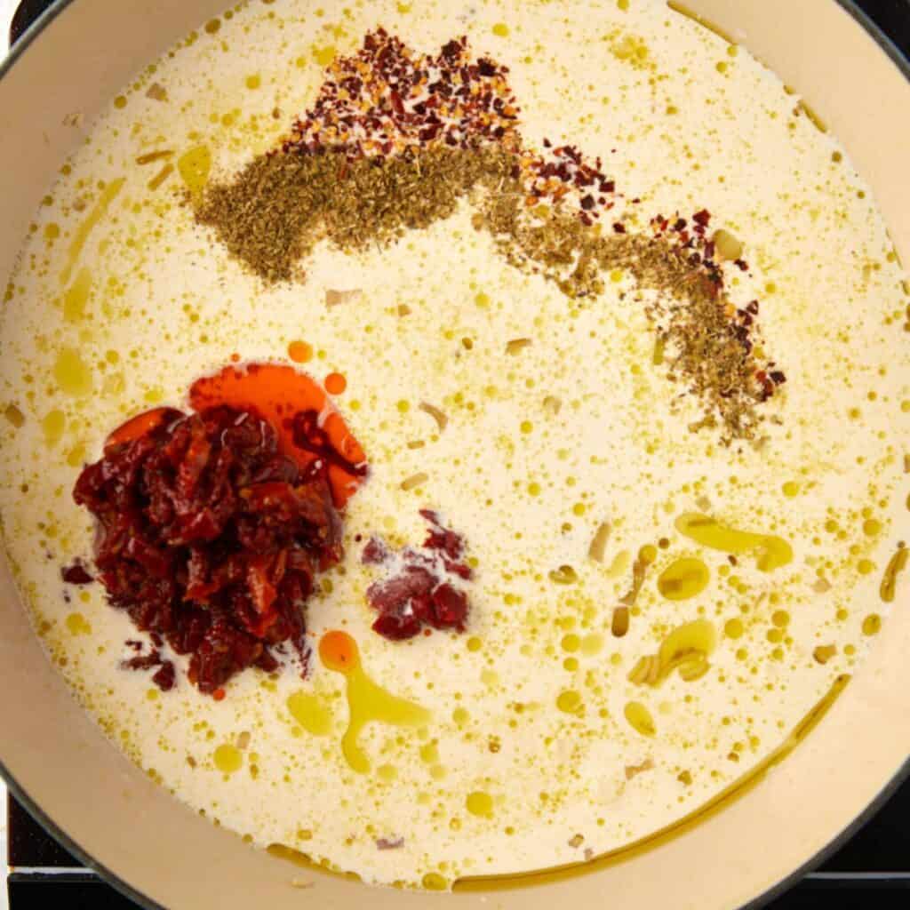 A creamy sauce in a pan with olive oil, dried herbs, crushed red pepper, and chopped sun-dried tomatoes, ready to be mixed together.