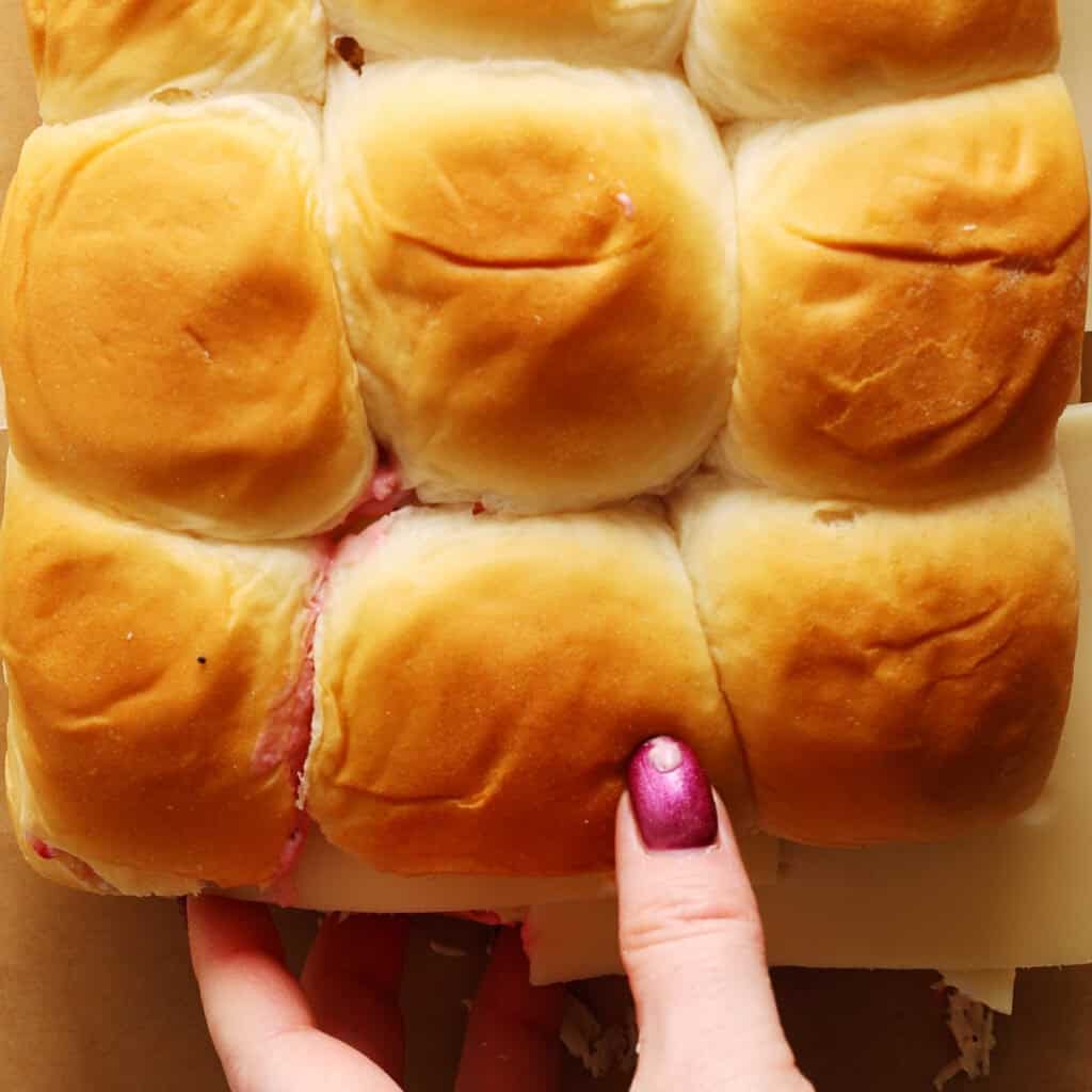 A hand with shiny purple nail polish holds a sheet of six golden, soft dinner rolls. Some faint filling and cheese are visible between the rolls.