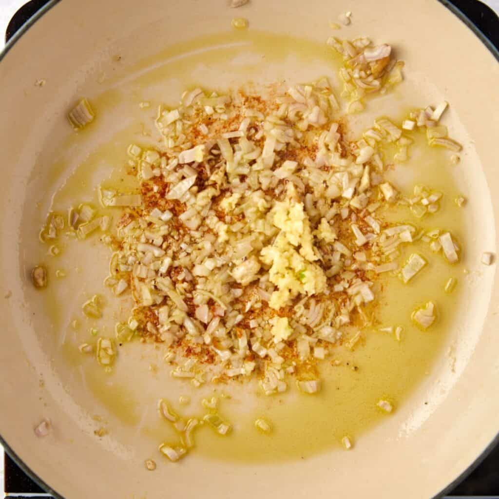 Chopped onions, garlic, and spices are sautéing in olive oil in a light-colored pan on a stovetop.