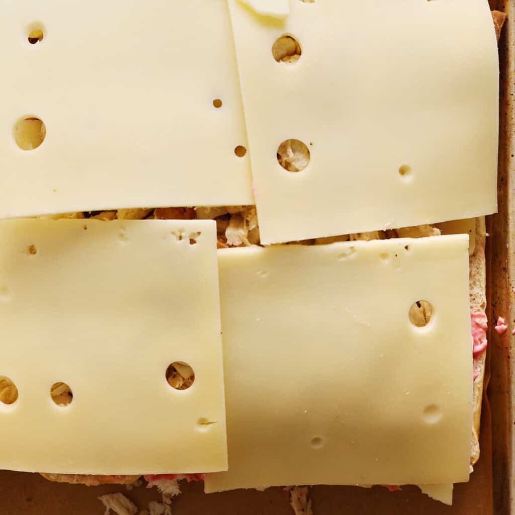 Close-up of several overlapping slices of Swiss cheese with holes placed on top of a sandwich with a light filling on brown bread.