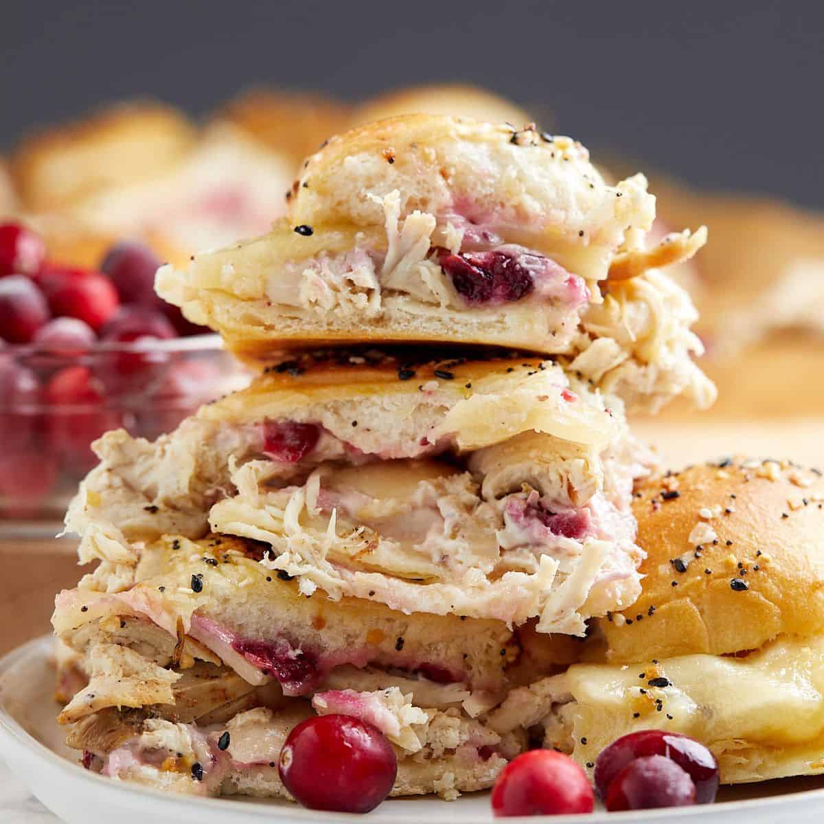 A close-up of stacked turkey and cranberry sliders on poppy seed buns, with melted cheese and a bowl of fresh cranberries in the background.