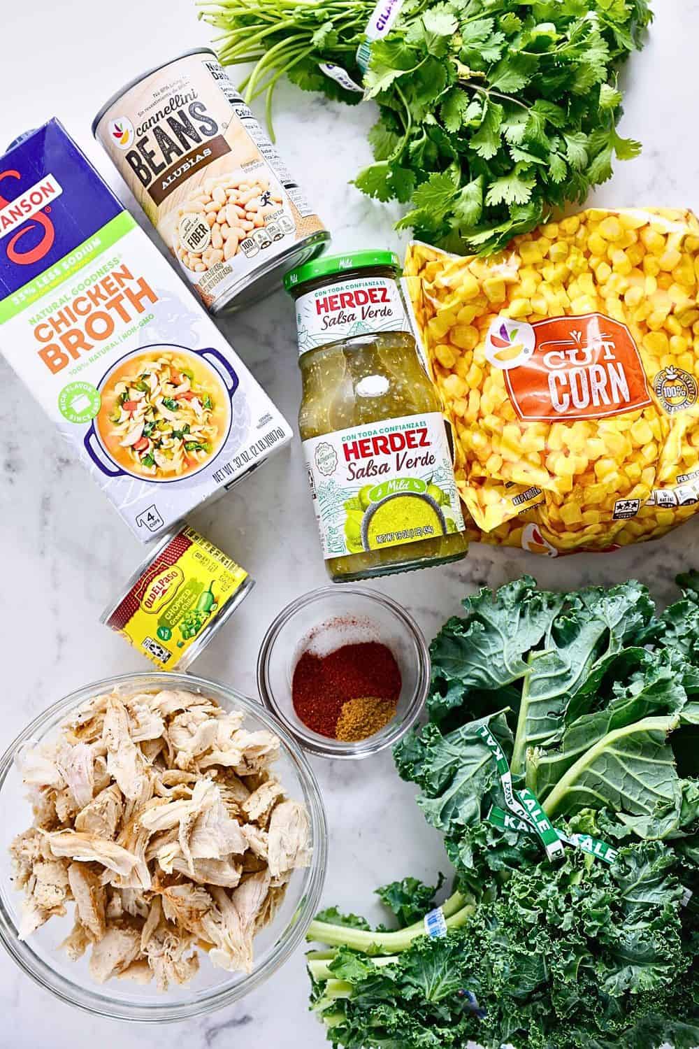 A flat lay of ingredients for a soup: chicken broth, canned cannellini beans, salsa verde, frozen corn, pulled chicken, green kale, cilantro, spices in a small bowl, and a bouillon cube on a white surface.