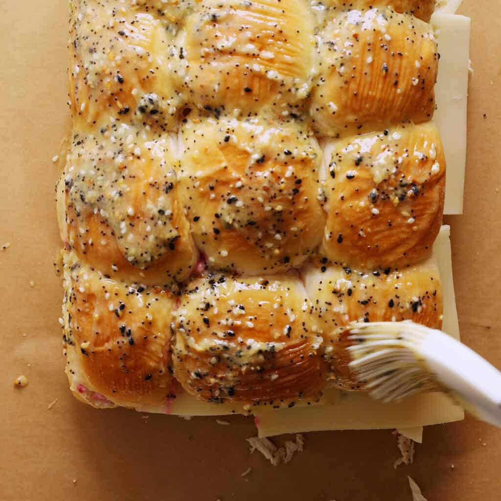 A close-up of a batch of sandwich sliders being brushed with a everything bagel seasoning 
mixture, showing shiny tops and visible cheese layers between the rolls on brown parchment paper.