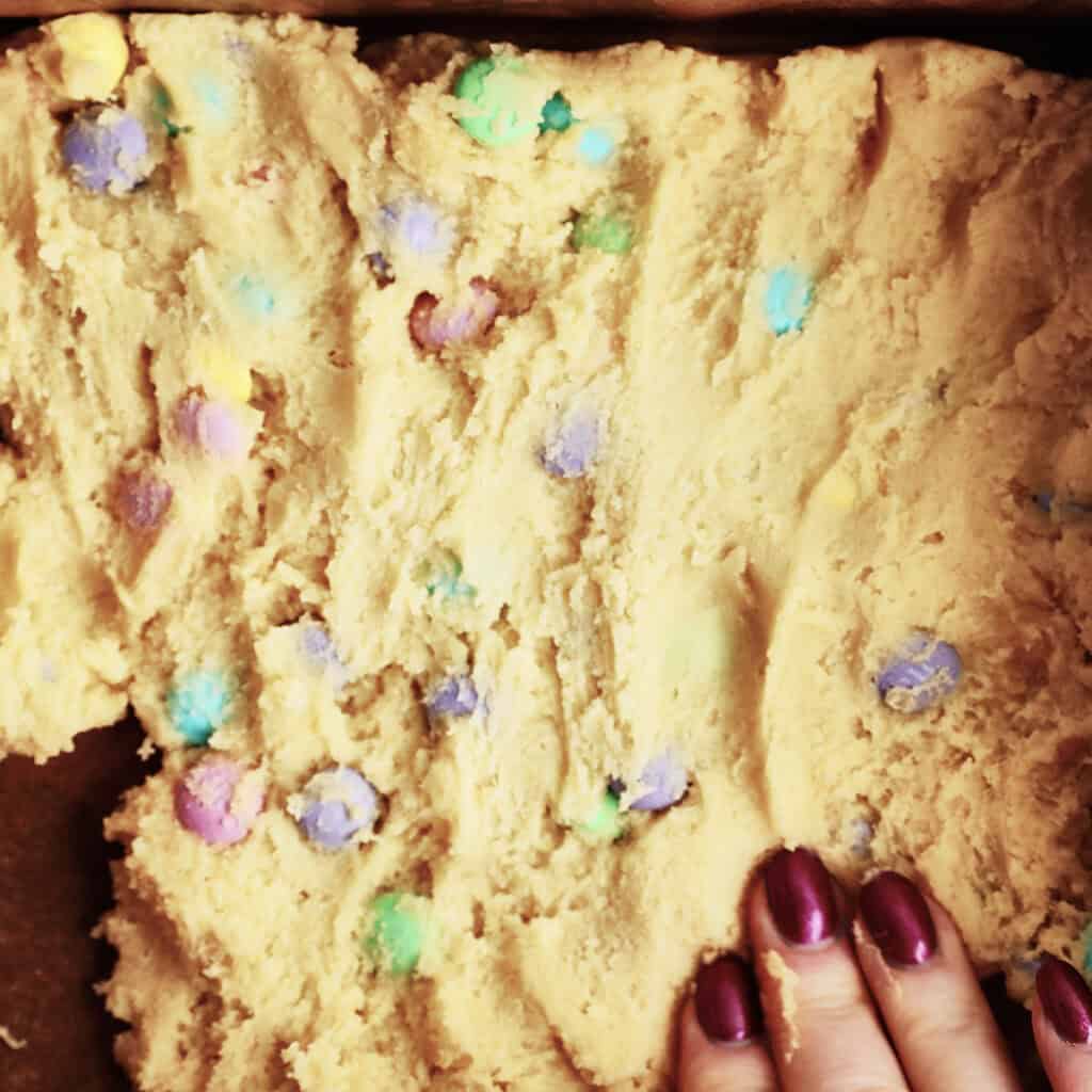 A hand with dark red nail polish presses cookie dough with colorful candy-coated chocolate pieces into a baking pan.