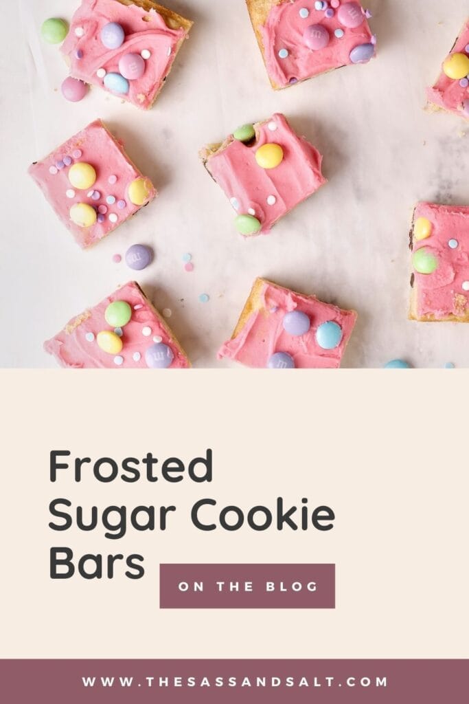 Several square sugar cookie bars topped with pink frosting and colorful candy-coated chocolates are arranged on parchment paper. Text below reads, "Frosted Sugar Cookie Bars – On the blog – thesassandsalt.com.