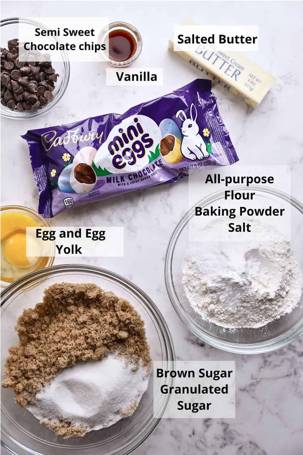 Baking ingredients on a white surface, including chocolate chips, a stick of butter, vanilla, an egg with yolk, a bag of Cadbury mini eggs, a flour mixture, and a bowl with brown and granulated sugar. Each item is labeled.
