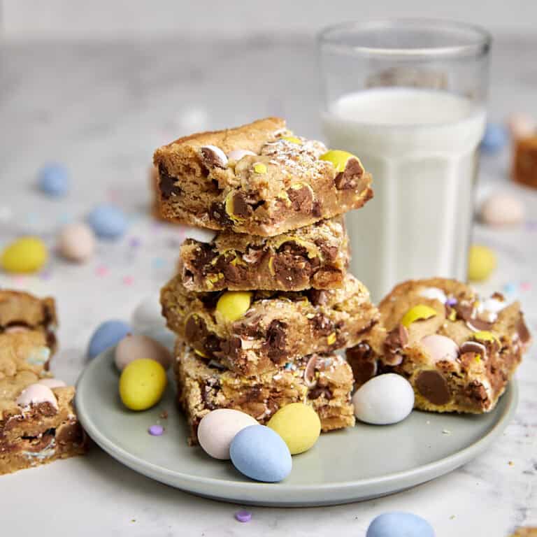A stack of blondie bars with chocolate candies and mini eggs on a gray plate, surrounded by pastel mini eggs, with a glass of milk in the background on a marble surface.