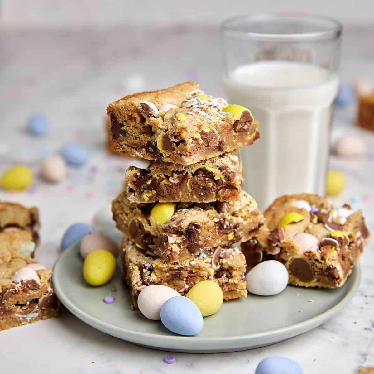 A stack of blondie bars with chocolate candies and mini eggs on a gray plate, surrounded by pastel mini eggs, with a glass of milk in the background on a marble surface.