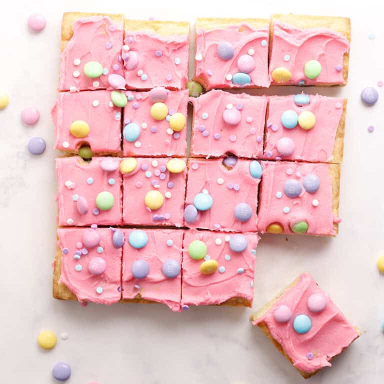 A batch of square sugar cookie bars topped with bright pink frosting, pastel-colored candy-coated chocolates, and sprinkles, arranged on a white surface. One bar is pulled slightly away from the group.