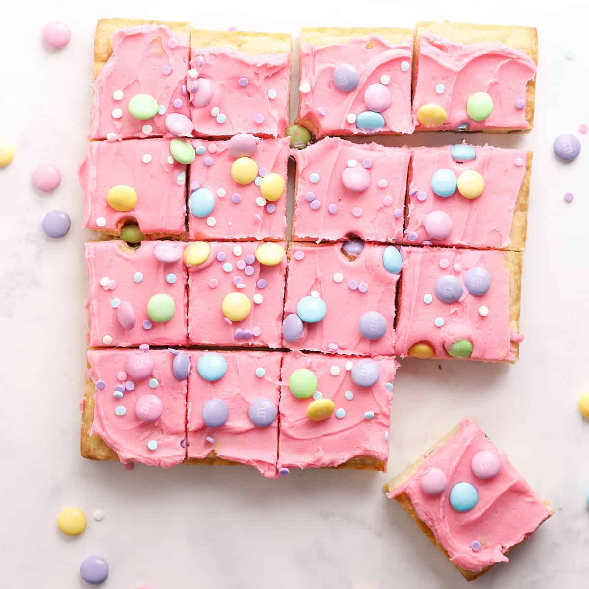 A batch of square sugar cookie bars topped with bright pink frosting, pastel-colored candy-coated chocolates, and sprinkles, arranged on a white surface. One bar is pulled slightly away from the group.