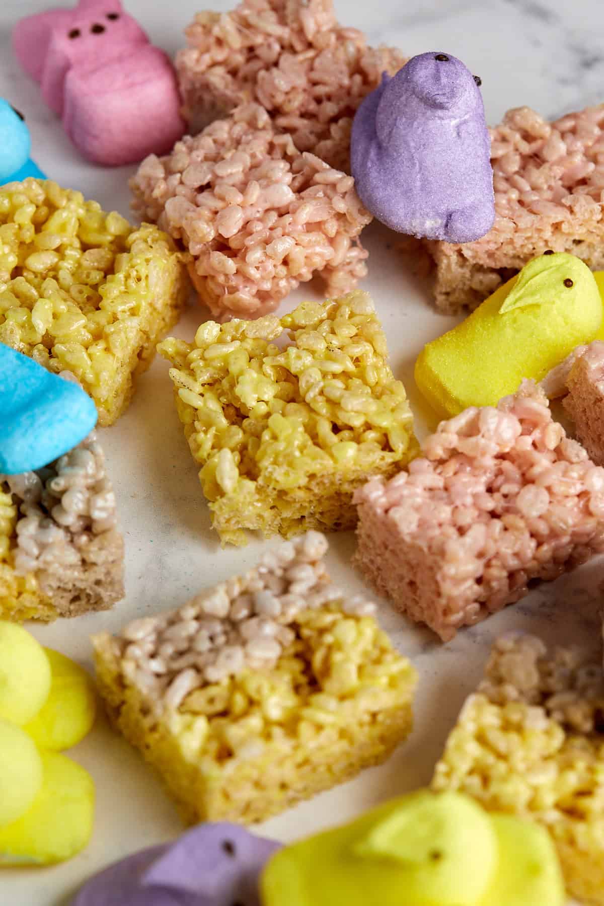 Marshmallow Peeps in yellow, blue, and purple are arranged with rice crispy treats, some cut into squares, on a white surface. The treats and candies create a colorful, festive display.