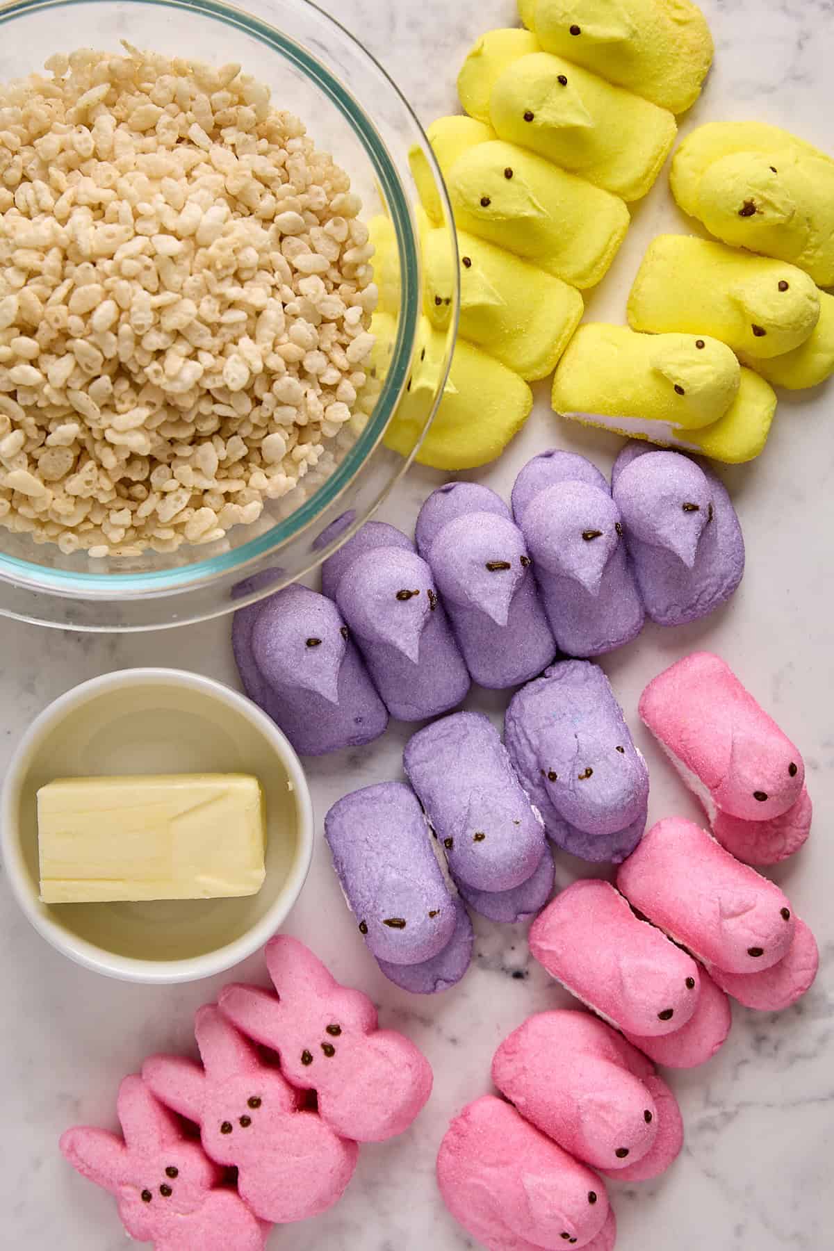 A bowl of crispy rice cereal, a small bowl of butter, and rows of marshmallow bunny Peeps in yellow, purple, and pink, all arranged on a marble surface, ready for making treats.