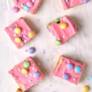 Five square sugar cookie bars with bright pink frosting, colorful candy-coated chocolates, and pastel sprinkles are arranged on white parchment paper. Some bars have bites taken out of them.