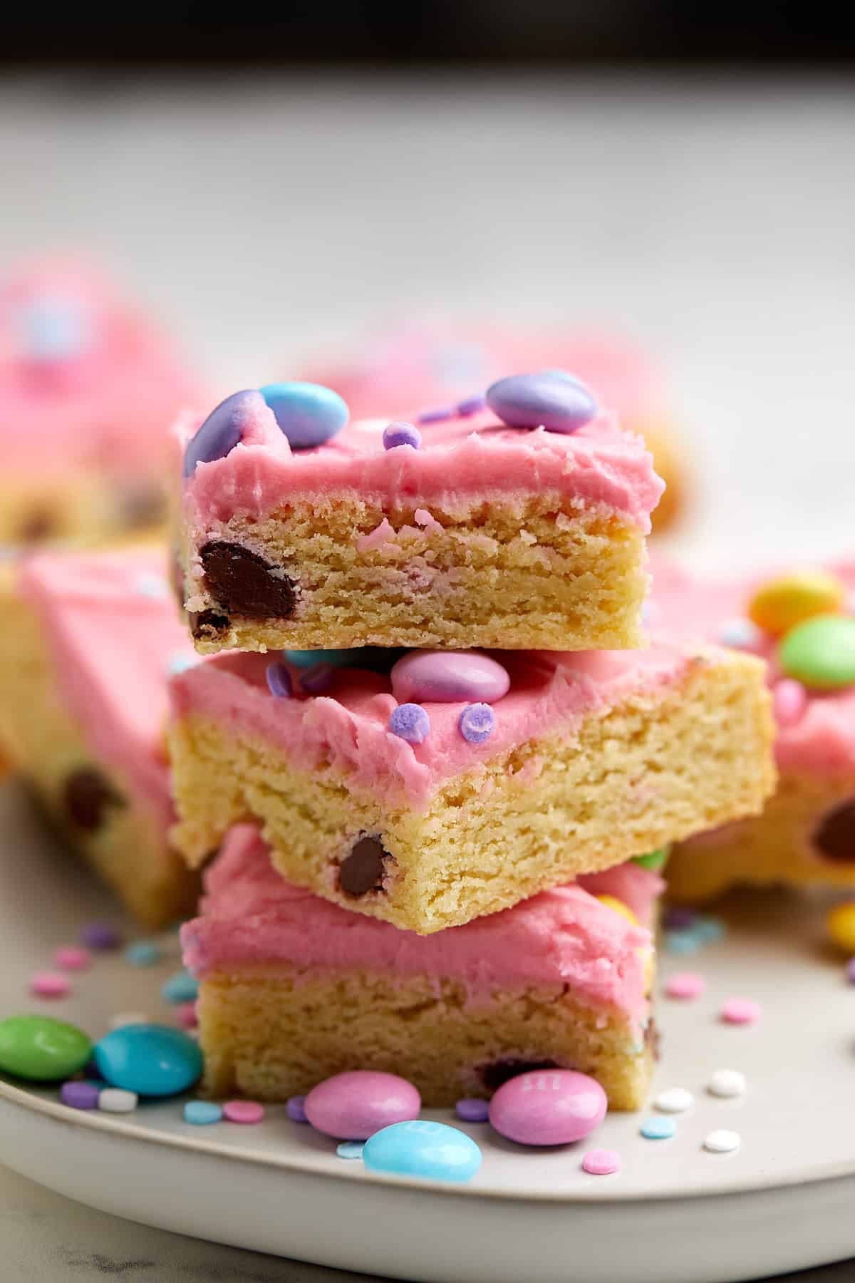 Three blondie bars with pink frosting and colorful candy-coated chocolates are stacked on a plate, surrounded by scattered candies and sprinkles. The bars have a soft, chewy texture with chocolate chips inside.