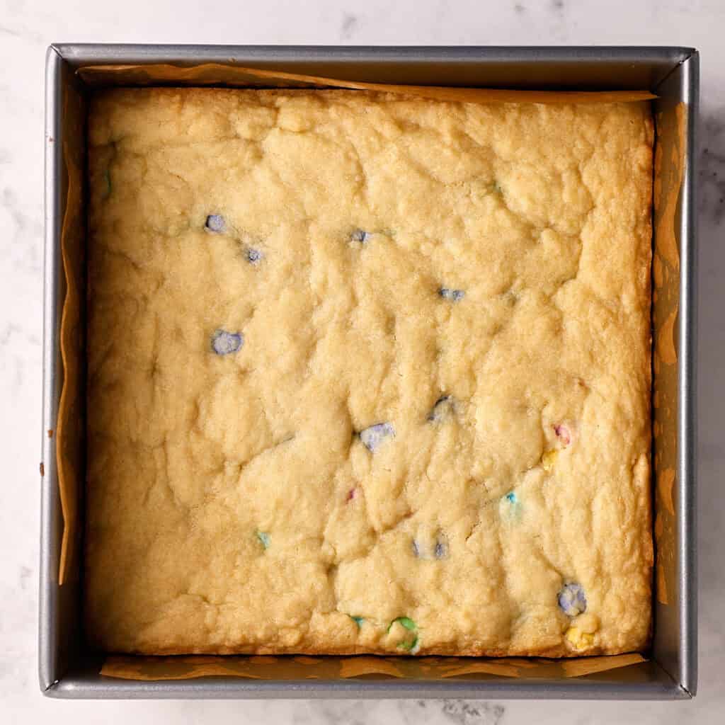 A square baking pan lined with parchment paper contains a large, golden-brown cookie bar with colorful candy pieces visible on the surface. The pan sits on a marble countertop.