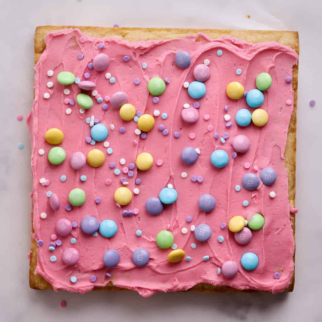 A square cookie with pink frosting, topped with pastel-colored candy-coated chocolates and round sprinkles, displayed on a white surface.