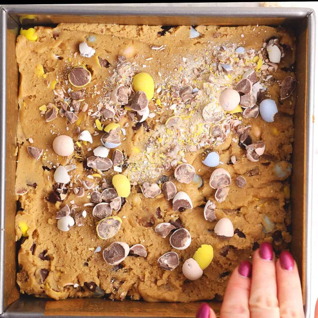 A square baking pan filled with cookie dough, topped with crushed and whole candy-coated chocolate eggs. A hand with painted nails holds the edge of the pan.