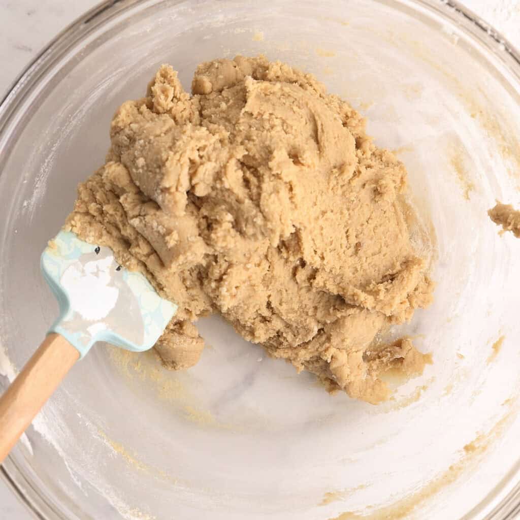 A glass mixing bowl containing thick, light brown cookie dough with a blue and white silicone spatula resting inside.