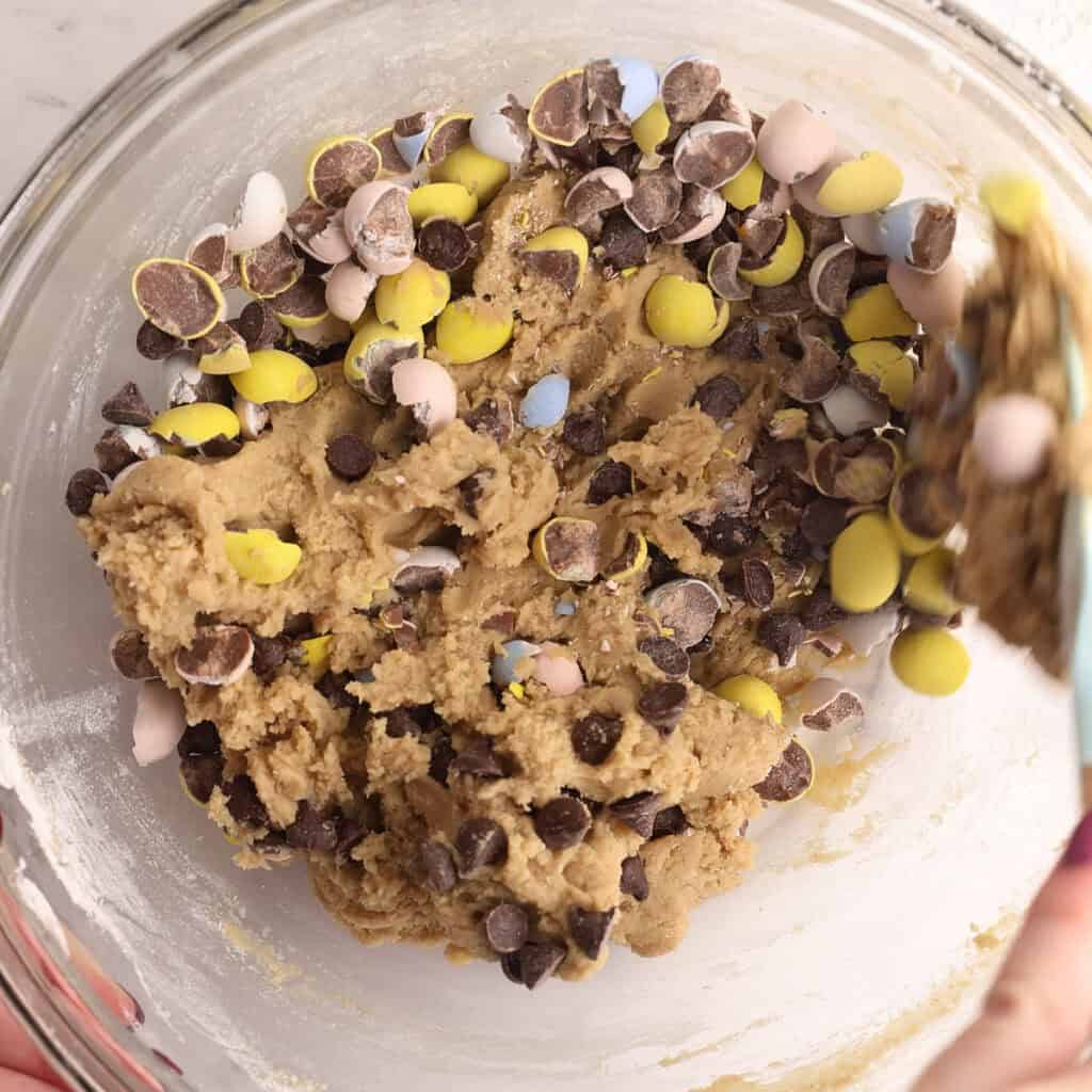 A glass bowl filled with cookie dough mixed with chocolate chips and colorful candy-coated chocolate eggs, being stirred with a spatula.
