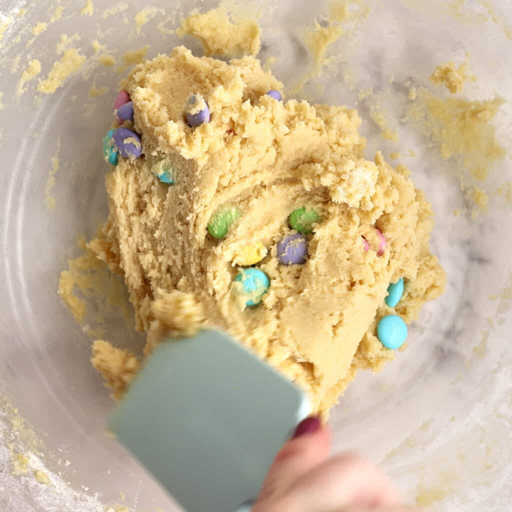 A hand holding a spatula mixes cookie dough with colorful candy-coated chocolates in a clear glass bowl.