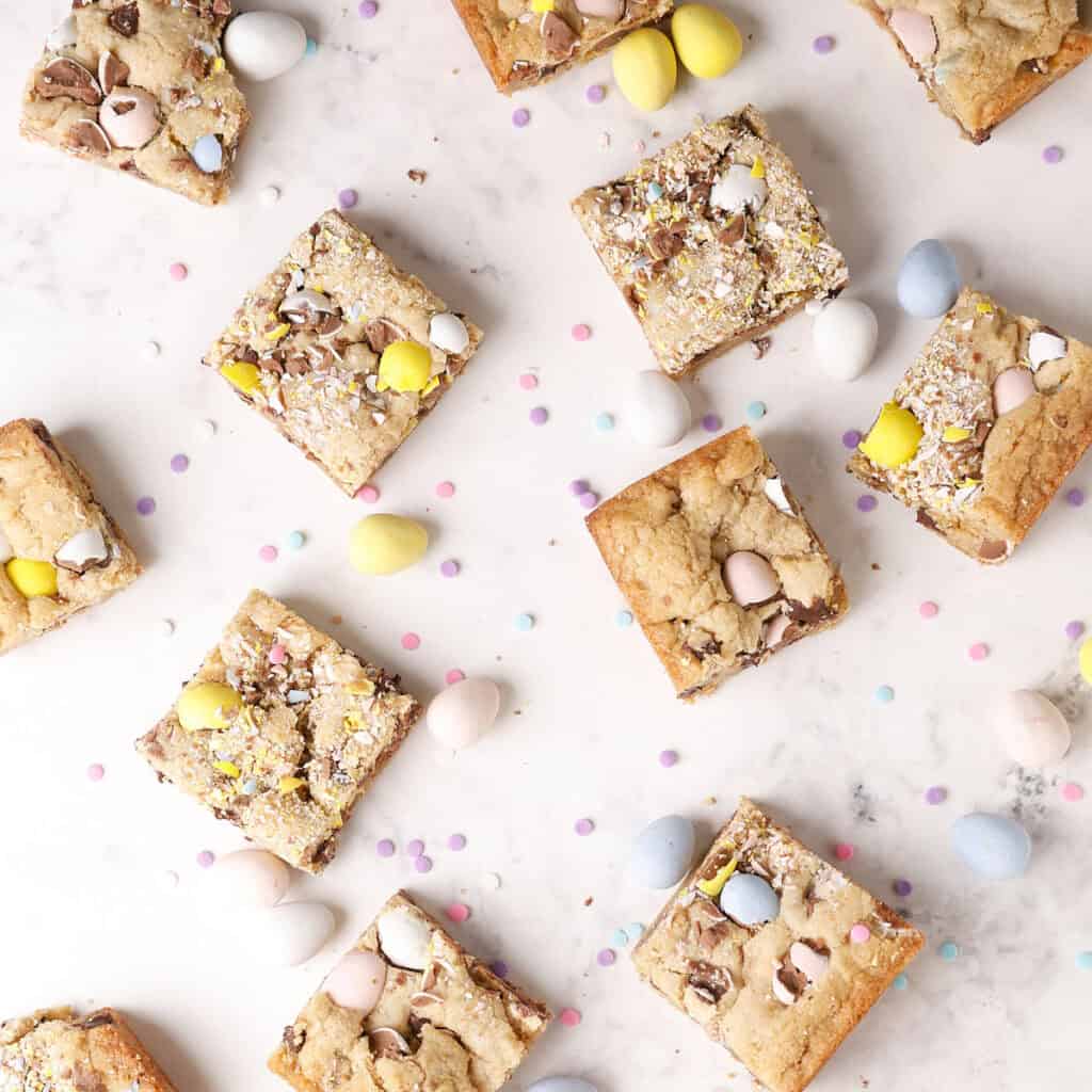 Top view of cookie bars with crushed chocolate eggs and mini candy eggs scattered on a white marble surface, along with small pastel confetti candies.