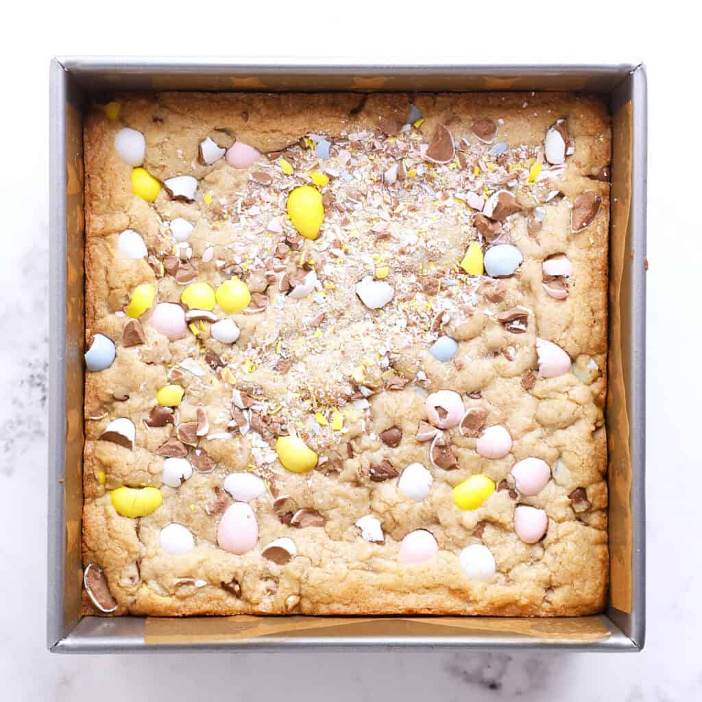 A square baking pan filled with a golden cookie bar topped with crushed pastel-colored chocolate eggs and sprinkles on a white background.