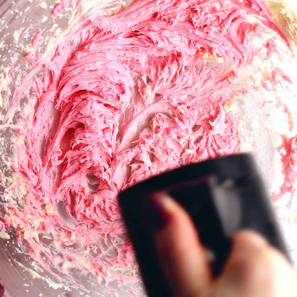 A close-up of a hand holding a black electric mixer blending bright pink frosting or batter in a mixing bowl. The swirled mixture looks creamy and thick.