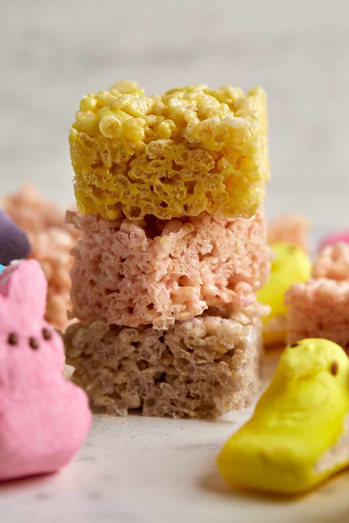 Three colorful rice crispy treats are stacked on top of each other, with yellow on top, pink in the middle, and brown at the bottom. Peeps marshmallow candies surround the stack.