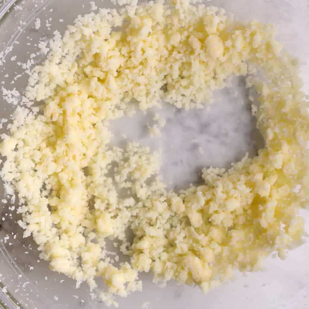 Small, pale yellow butter granules clumped together in a clear mixing bowl, with a ring-shaped gap in the center showing the bottom of the bowl.