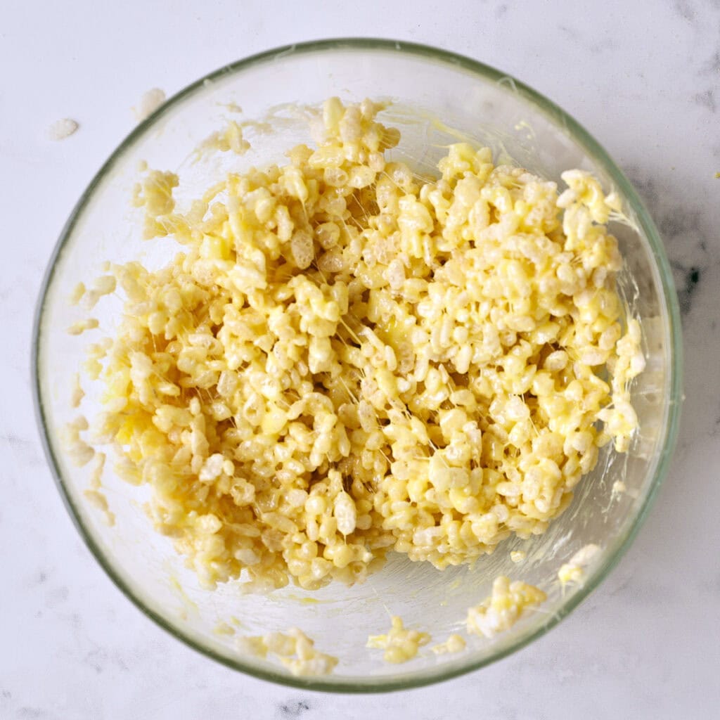 A glass bowl filled with a mixture of Rice Krispies cereal coated in a creamy, melted marshmallow mixture, sitting on a white marble surface.