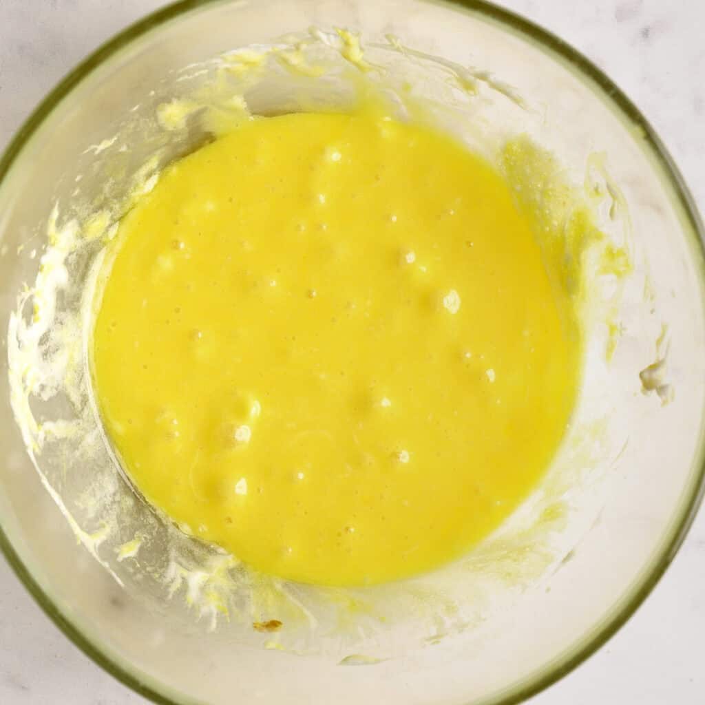 A glass bowl containing a bright yellow, slightly lumpy batter or mixture, set on a light-colored surface.