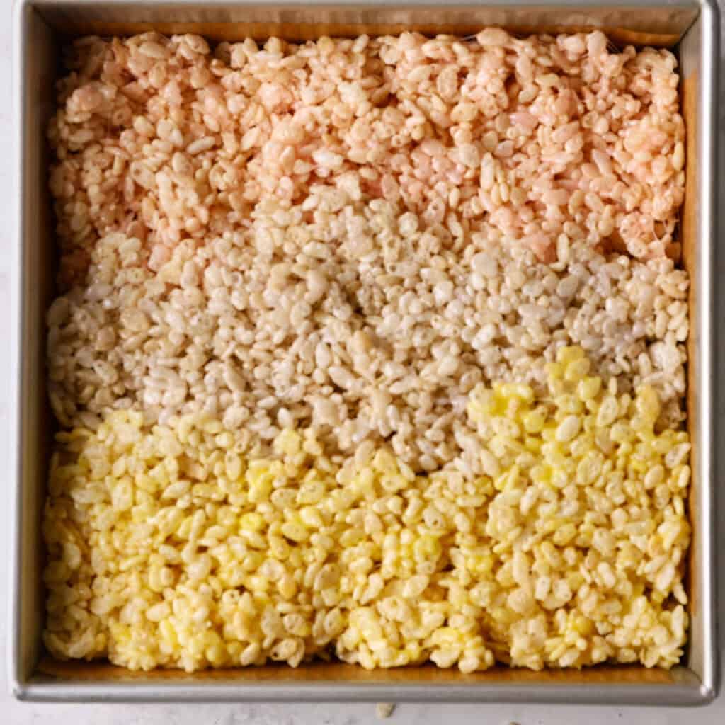 A square baking pan filled with Rice Krispies treats arranged in three horizontal layers: pink on top, beige in the middle, and yellow on the bottom.
