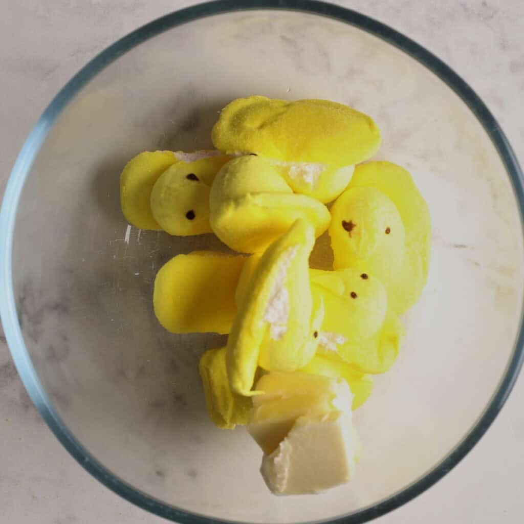 A glass bowl containing several yellow marshmallow chicks and a chunk of butter, placed on a white marble surface.