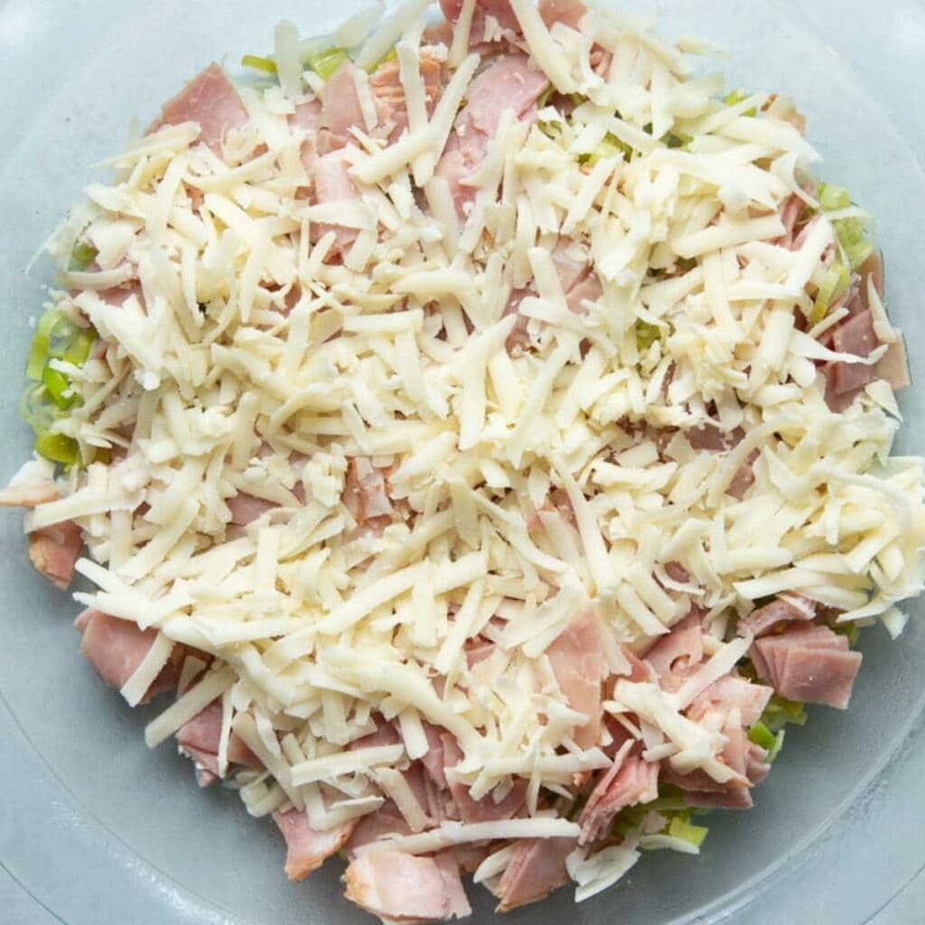 Sautéed leeks, diced ham, and shredded Gruyère layered in a glass pie dish, ready for the custard.
