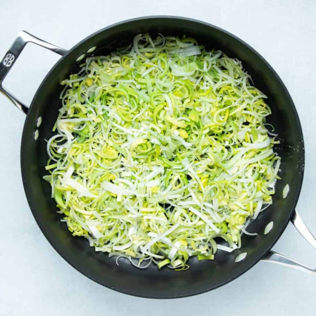 Sliced leeks sautéing in butter in a black skillet until soft and silky the first step in a crustless leek quiche.
