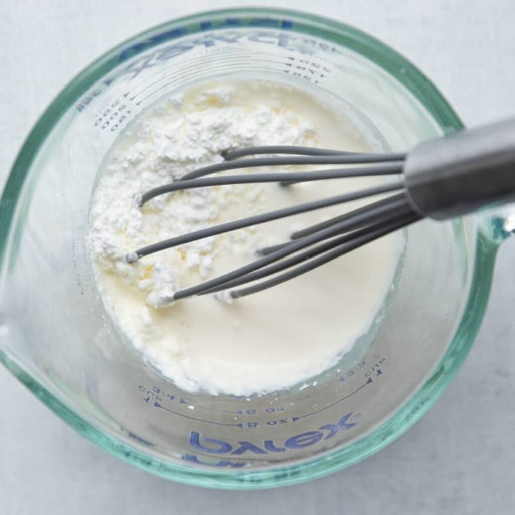 A whisk stirring cornstarch into cold milk in a Pyrex measuring cup to make the slurry for a silky quiche custard.