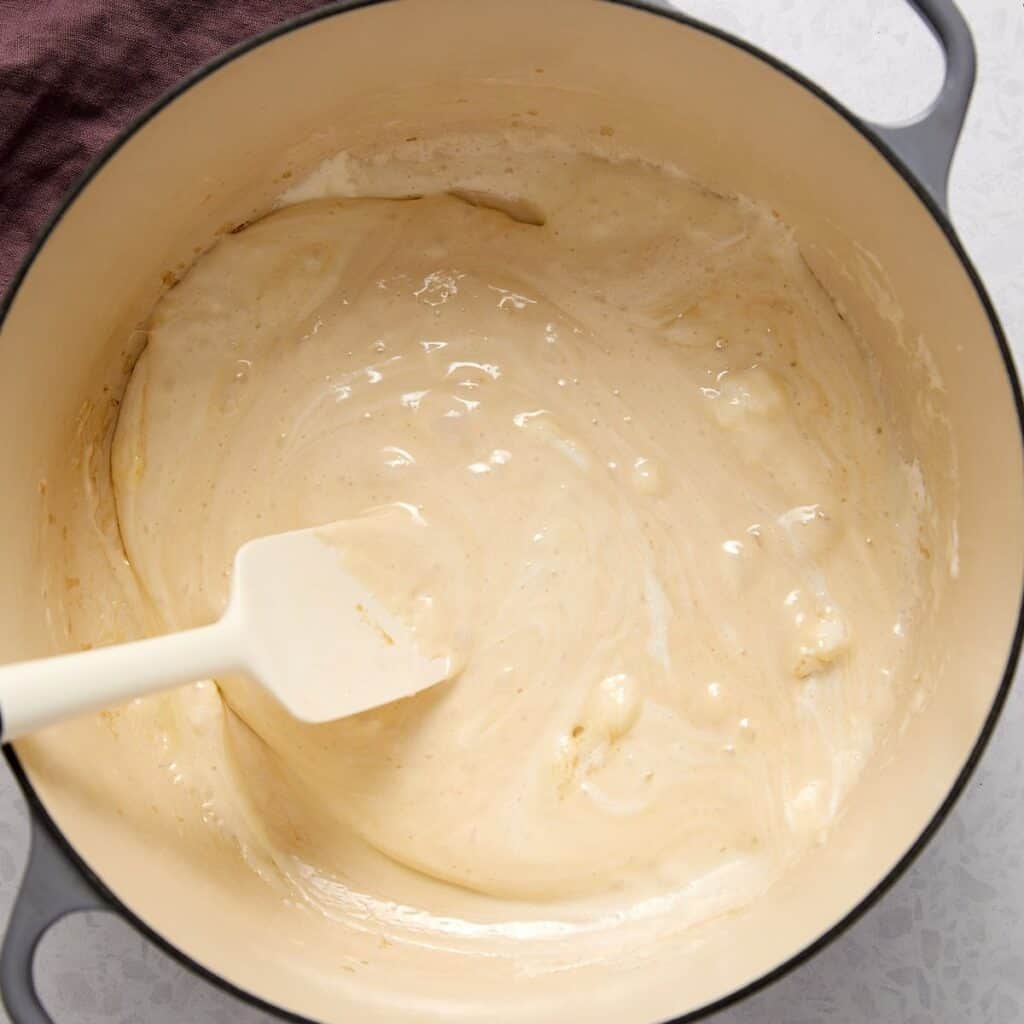 A creamy, light beige mixture being stirred with a white spatula in a large, light-colored pot. The mixture appears smooth with some lumps.